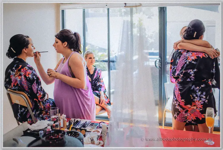 Bridesmaids in floral robes have makeup applied near glass doors as morning light fills the room.