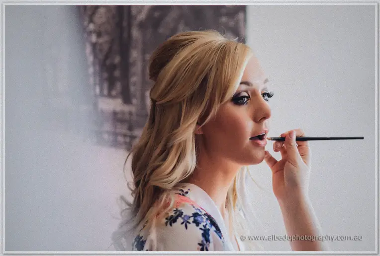 A makeup artist touches up blonde bride Kristyn's lipstick in soft sidelight before the ceremony.