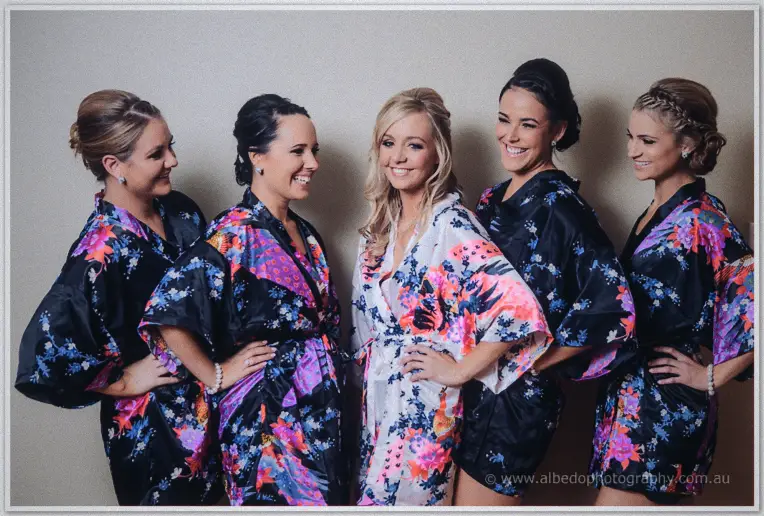 Kristyn and her bridesmaids beam in matching floral robes, hands on hips against a pale wall.