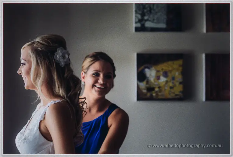 A bridesmaid in cobalt blue laughs behind Kristyn as her curls are finished beside framed art.