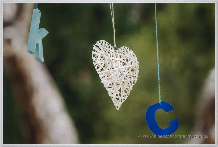 A woven rattan heart hangs between blue letters K and C on strings from a garden tree.