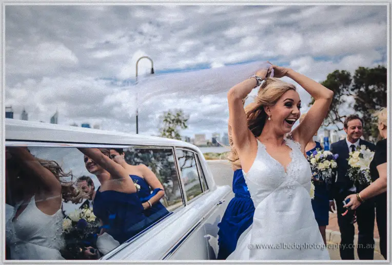 Kristyn throws her head back laughing, veil aloft, reflected in the limousine window with bridesmaids.