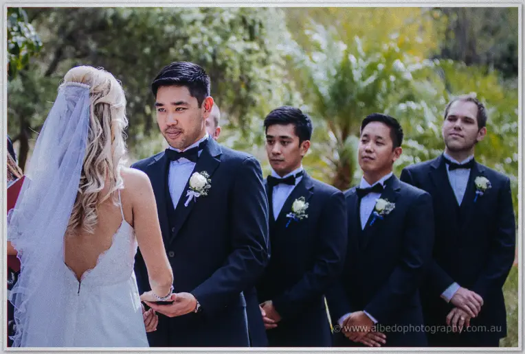 Chris holds Kristyn's hands and gazes at her while groomsmen line up under soft garden light.
