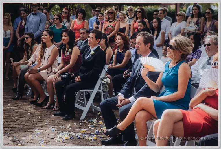 Wedding guests on white chairs fan themselves and smile as petals scatter across the brick aisle.