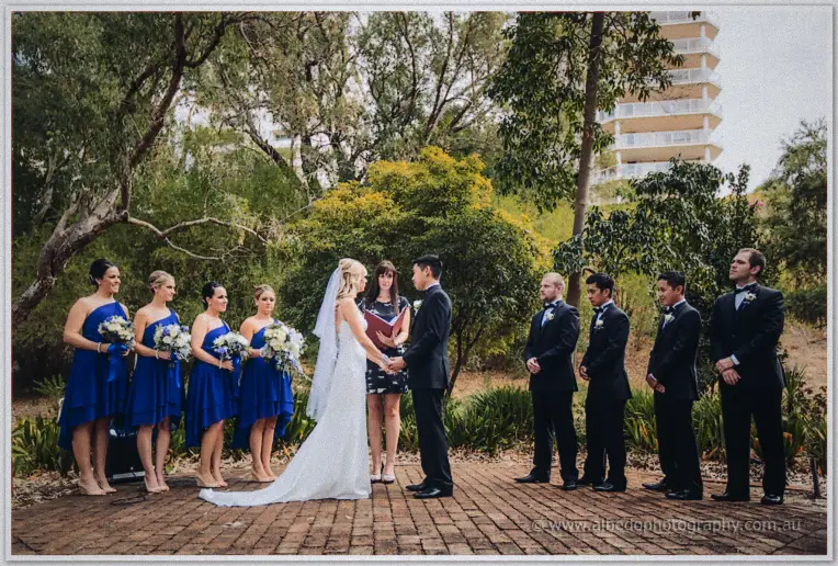 Kristyn and Chris hold hands before the celebrant, flanked by blue-dressed bridesmaids and tuxedoed groomsmen.