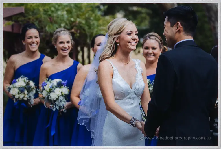 Kristyn beams at Chris mid-ceremony as bridesmaids in cobalt smile behind them among the trees.