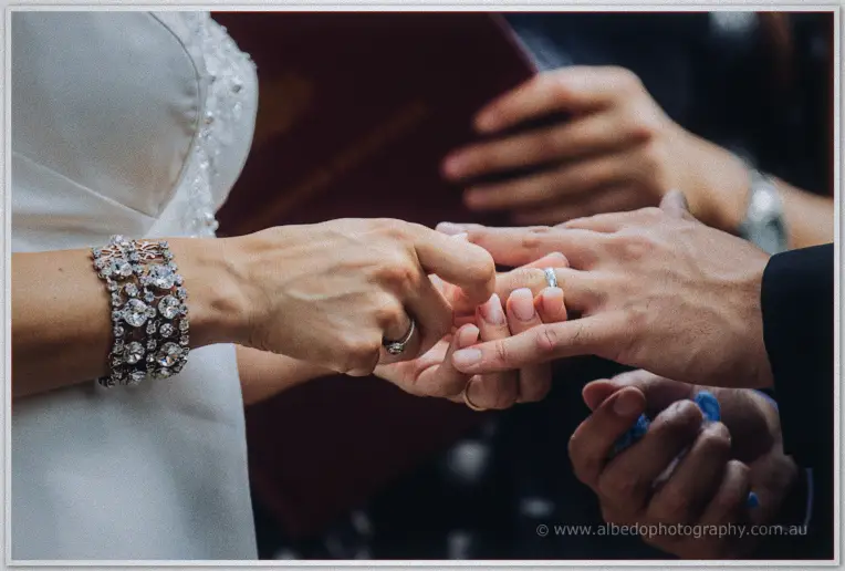 Close-up of Kristyn sliding the wedding ring onto Chris's finger, her jewelled bracelet catching light.