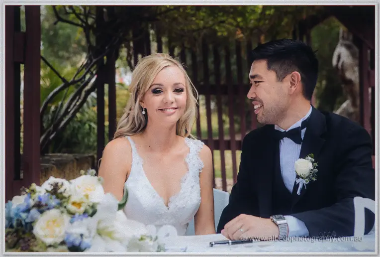 Kristyn and Chris smile at the signing table beneath a maroon pergola, bouquet of white roses beside them.