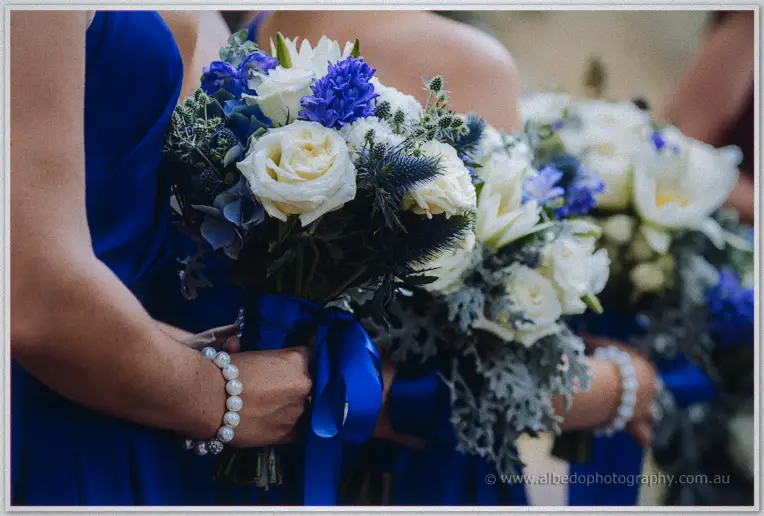 Bridesmaids clasp bouquets of white roses, blue hyacinths and thistles tied with royal blue ribbon.