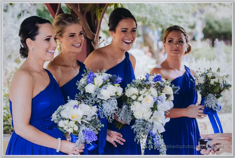 Four bridesmaids in one-shoulder cobalt gowns laugh while holding blue and white bouquets in the garden.