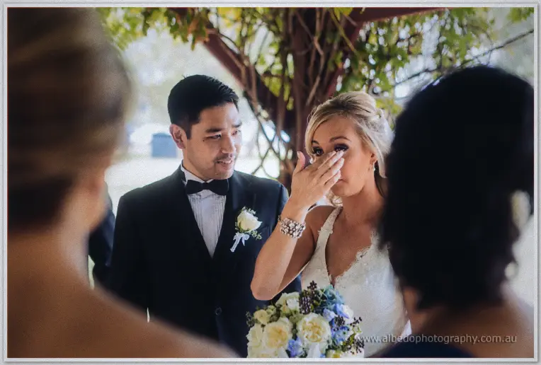 Kristyn wipes away a tear as Chris smiles at her beneath the pergola, guests soft in the foreground.