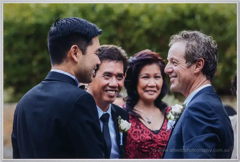 Chris greets smiling wedding guests in suits and a red lace dress outside the garden ceremony.
