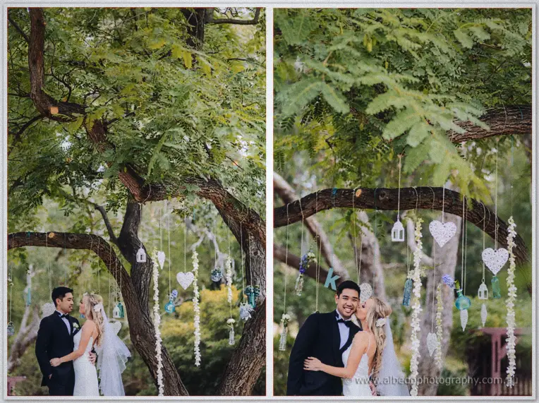 The couple embrace beneath a tree hung with lace hearts, glass lanterns and dangling white blooms.