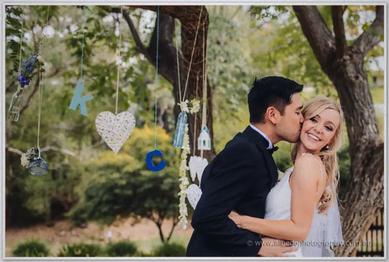 Chris kisses Kristyn's cheek beneath hanging wicker hearts and blue letters K and C in the garden.