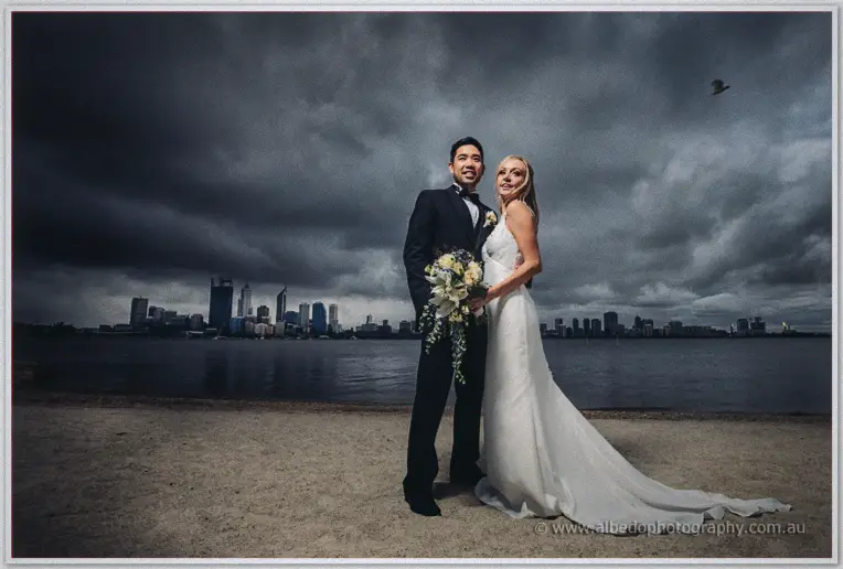Kristyn and Chris stand on the river foreshore, Perth skyline rising behind dramatic storm clouds.