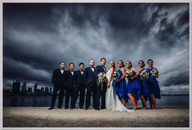 The full wedding party lines the sandy riverbank under heavy skies, Perth towers behind them.