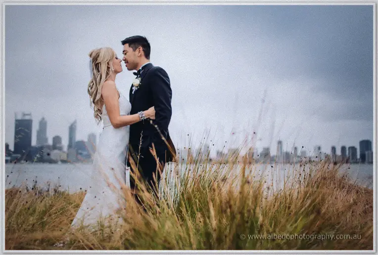Kristyn and Chris lean in for a kiss on the South Perth foreshore, the city skyline hazy across the water