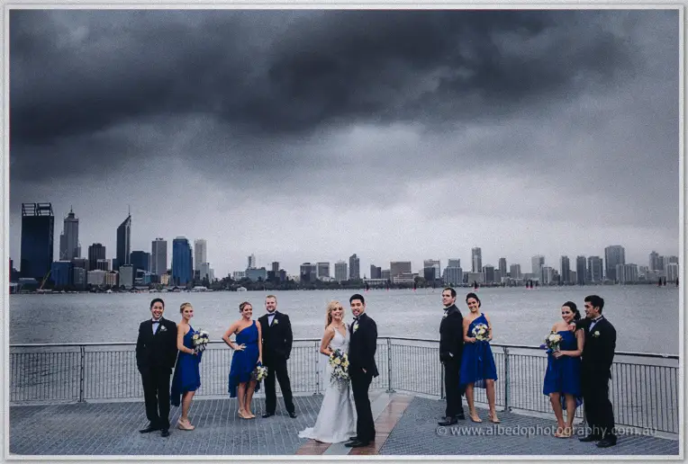 Bridal party in cobalt dresses and dark suits line the waterfront rail beneath the stormy city skyline