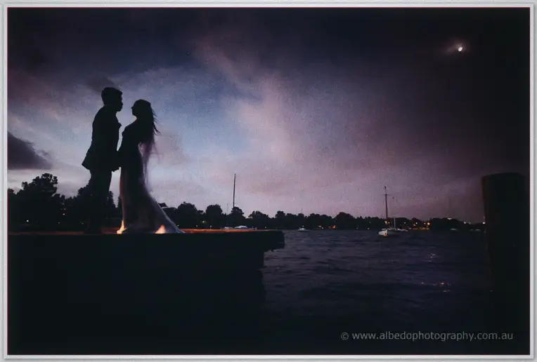 Silhouetted against a violet twilight sky, the newlyweds stand close together on the jetty above moored boats