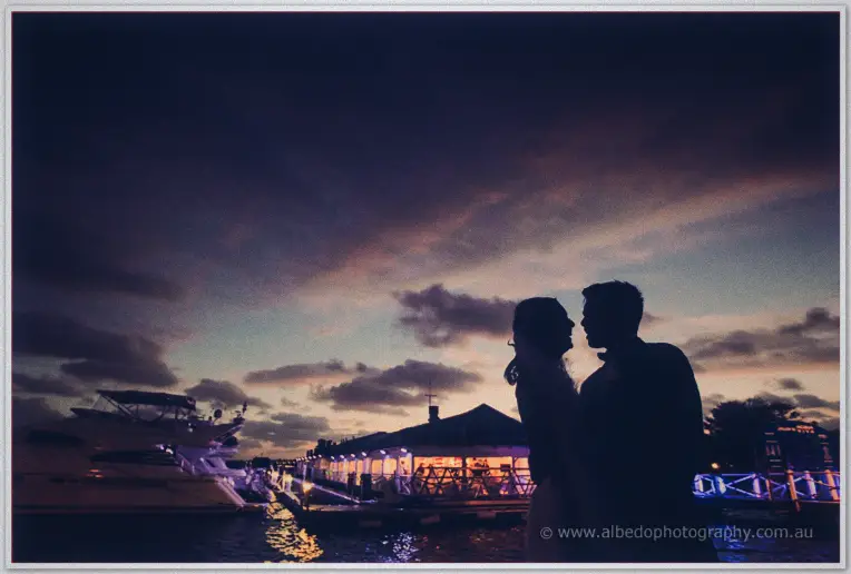The couple stand silhouetted at dusk on the jetty, warm pavilion lights glowing behind moored yachts