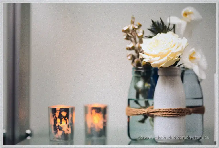 A cream rose in a twine-wrapped bottle stands among blue glassware and softly glowing votive candles