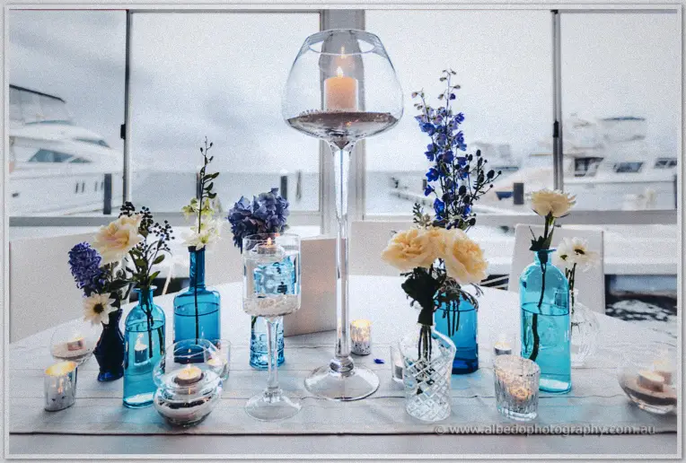 Blue bottles of roses and hydrangeas crowd the table before windows framing yachts in the marina