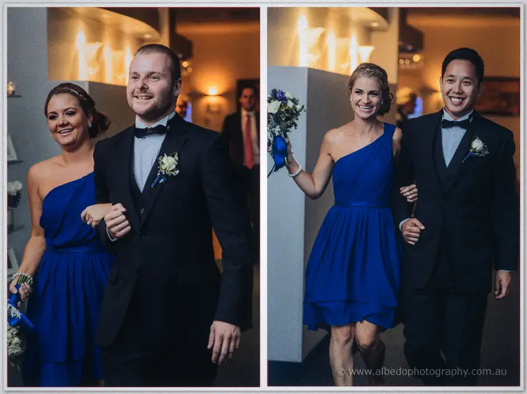 Two more bridal party pairs enter laughing, blue bouquets swinging past the softly glowing wall sconces