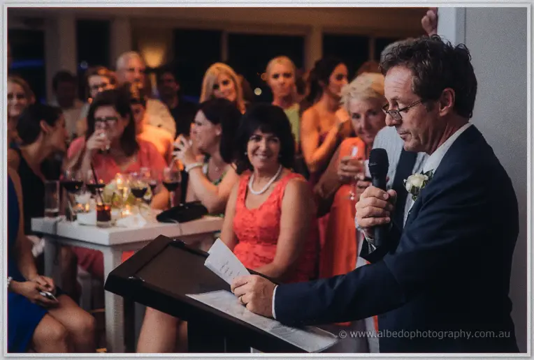 A suited guest speaks at the lectern while wedding guests crowd the candlelit tables listening closely