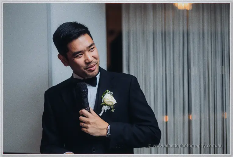 Chris grins into the microphone mid-speech, white rose boutonniere bright against his black tuxedo