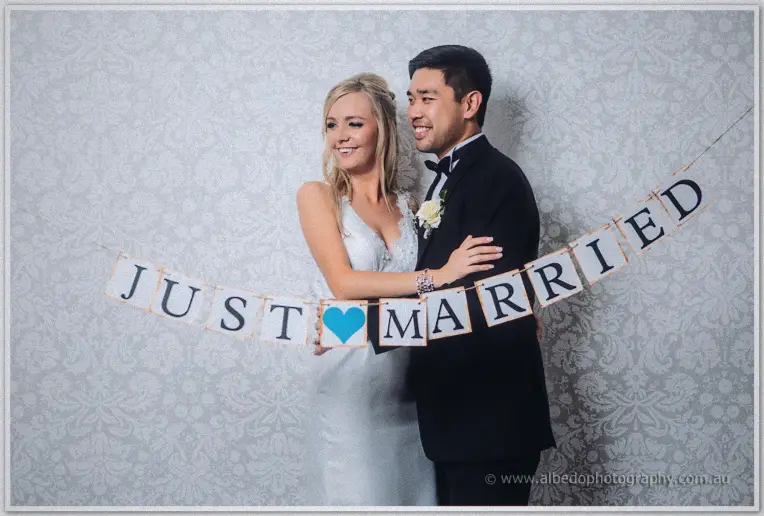Behind a just married bunting with a blue heart, Kristyn and Chris embrace against the damask backdrop