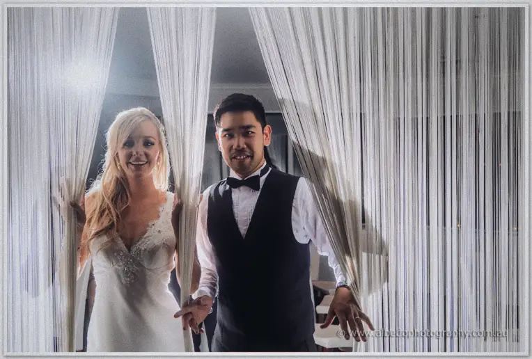 Kristyn and Chris peek playfully through parted strands of a white string curtain, backlit and grinning
