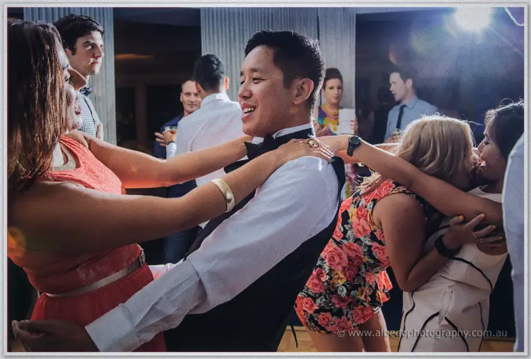 A packed dance floor swirls around a smiling Chris in black vest amid guests in coral and floral dresses
