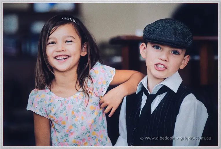 A flower girl loops her arm around the page boy as both giggle in a lamplit lounge room
