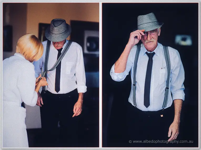 An older gentleman in braces and a grey trilby has his tie clipped by a blonde helper, then tips his hat