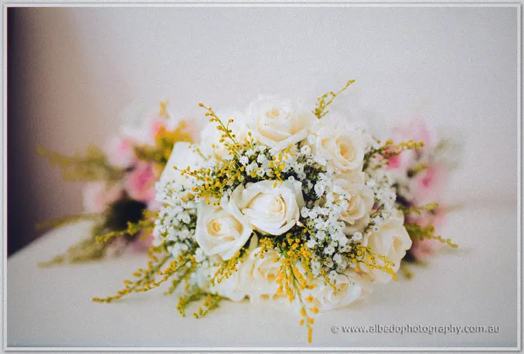 A cream rose and wattle bridal bouquet with baby's breath rests on a table beside pastel posies