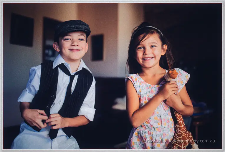 The page boy buttoning his pinstripe vest and the flower girl hugging a toy giraffe both grin widely