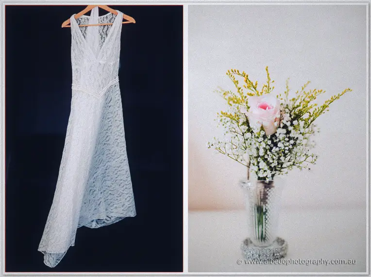 The lace bridal gown hangs on a hanger against navy while a single pink rose brightens a crystal vase