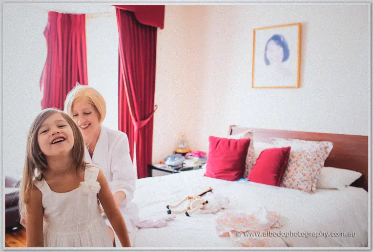 A blonde bridesmaid laughs with the flower girl beside red-curtained windows in a bright bedroom
