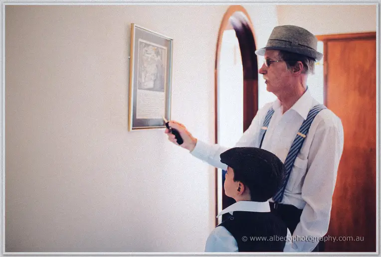 A bespectacled groomsman in suspenders points at a framed etching while the page boy watches