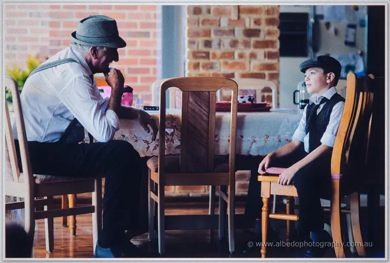 An older groomsman in a trilby and the page boy sit apart at a kitchen table chatting before the ceremony