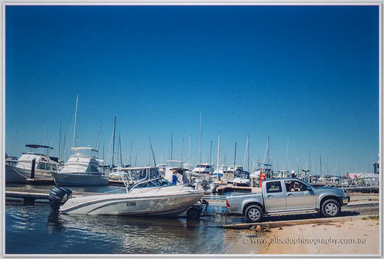 A ute launches a tinnie at the sandy ramp beside moored yachts under a cloudless blue sky