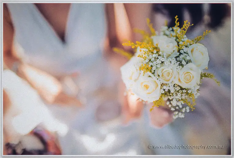 Tansy holds her white rose and wattle bouquet aloft against the sunlit sky at the marina