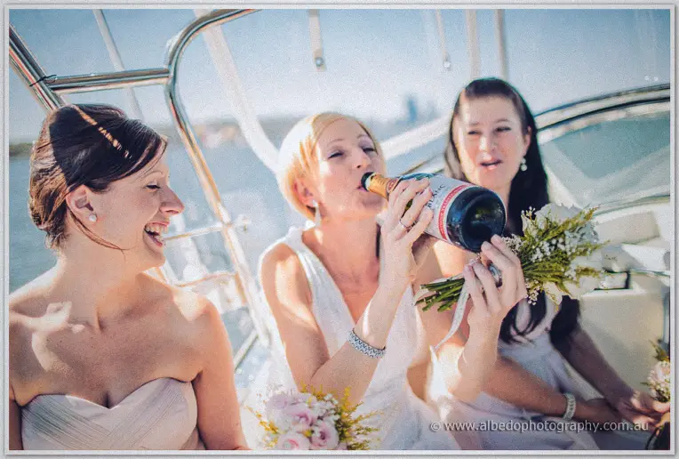 Tansy swigs champagne from the bottle between two laughing bridesmaids aboard the boat