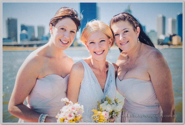 Tansy beams between her two bridesmaids in blush gowns with the city skyline across the river