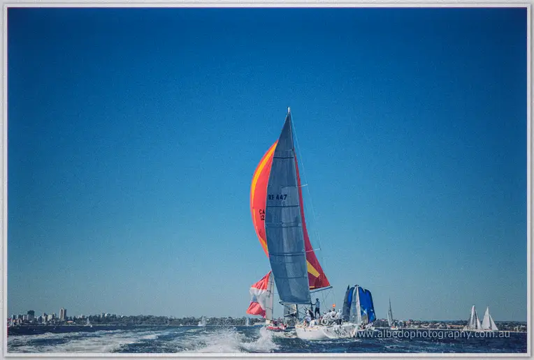 Yachts with red and grey spinnakers heel across the river beneath a towering cobalt sky