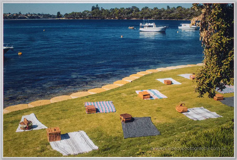 Picnic rugs and wicker baskets line the grassy foreshore beside moored boats at Green Place Reserve
