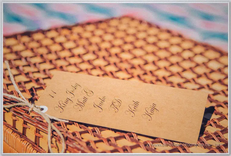 A handwritten kraft guest tag tied with twine lies across the lid of a wicker picnic basket