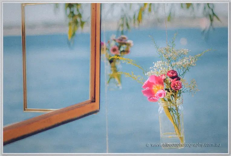 A hanging jar of pink blooms and wattle sways beside a gilt frame against the river haze