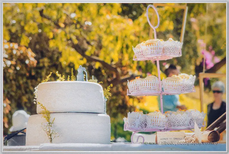 A two-tier white wedding cake stands beside a pink stand of cupcakes in dappled garden light
