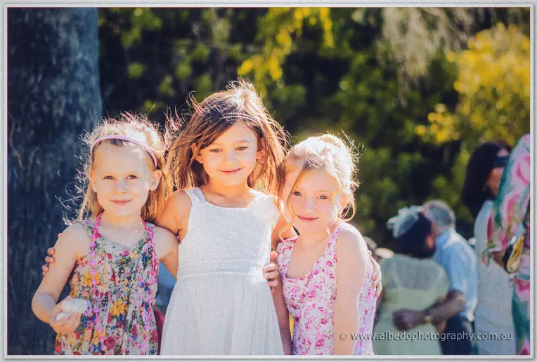 Three young girls in floral frocks and one white dress huddle arm in arm in golden afternoon light